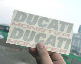 Puede incluir: Dos pegatinas Ducati Monster se sostienen contra un fondo borroso al aire libre. Las pegatinas son rectangulares y presentan las palabras "DUCATI" y "MONSTER" en una combinación de colores gris y rojo. Las pegatinas son sostenidas por una mano.