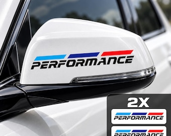 Puede incluir: Un espejo lateral de coche blanco con una calcomanía que muestra la palabra PERFORMANCE en letras mayúsculas negras, situada bajo tres franjas horizontales en azul claro, azul oscuro y rojo. El set incluye dos calcomanías idénticas.