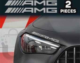 Puede incluir: Dos calcomanías plateadas AMG con un diseño de rayas inclinadas. El conjunto incluye dos piezas, como indica el texto. Una calcomanía se muestra aplicada en el capó de un coche negro mate, sobre el conjunto del faro.