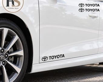 Puede incluir: Un juego de dos pegatinas de vinilo negro con el logotipo de Toyota y la palabra TOYOTA en una fuente sans-serif. Las pegatinas están aplicadas en el panel lateral inferior de un coche blanco, con texto adicional visible en la esquina superior.