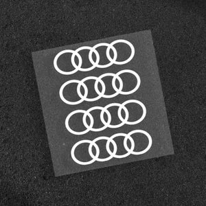 May include: A sheet of white Audi logo decals. The decals are arranged in four rows of interlocking rings, a classic automotive emblem. The decals are on a black background.