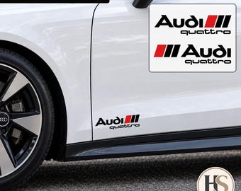 Puede incluir: Un juego de 2 pegatinas de vinilo Audi quattro en negro y rojo aplicadas en el lateral de un coche blanco. Los adhesivos muestran el texto del logotipo de Audi con un elemento gráfico de rayas rojas y negras. El set incluye dos pegatinas iguales.
