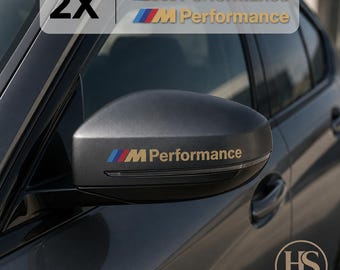 Puede incluir: Un juego de dos pegatinas M Performance con las icónicas franjas azul, morada y roja junto al texto M Performance en dorado. Las pegatinas están aplicadas sobre la carcasa del retrovisor negro mate de un vehículo de color oscuro.