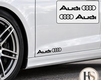 Puede incluir: Un juego de 2 pegatinas de vinilo negro para coche con el nombre de la marca Audi y el logotipo de los cuatro aros. Las pegatinas se muestran aplicadas en el panel lateral inferior de un vehículo blanco, junto a una imagen de ambas.