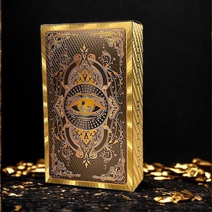 Puede incluir: Una caja de naipes dorada y marrón con diseños ornamentados y el texto "GOLD FOIL". La parte delantera presenta un símbolo de ojo detallado. La caja es rectangular y tiene un aspecto texturizado, con detalles dorados.