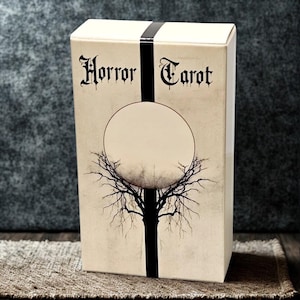 Peut inclure: Une bo&icirc;te de cartes de tarot beige avec les mots "Horror Tarot" en police gothique. Une bande noire verticale traverse le devant, croisant un cercle blanc avec une illustration d'arbre. La bo&icirc;te est sur une surface marron.