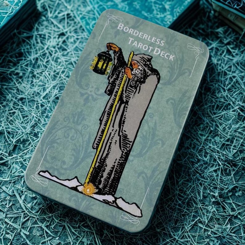 Peut inclure: Bo&icirc;te rectangulaire pour jeu de tarot avec un fond turquoise et l'inscription "BORDERLESS TAROT DECK" en haut. L'illustration repr&eacute;sente une figure v&ecirc;tue d'une robe tenant une lanterne et un b&acirc;ton. La bo&icirc;te a un motif floral d&eacute;coratif.