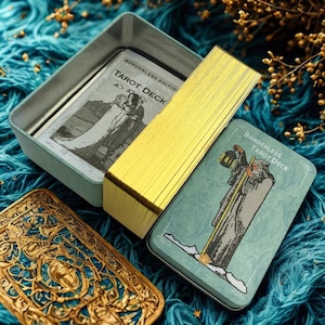 Può includere: Un mazzo di tarocchi con bordi dorati e una scatola di latta color verde acqua. La scatola reca la scritta "Borderless Tarot Deck" e presenta illustrazioni di figure. Il mazzo è aperto, rivelando le carte e il contenuto della scatola.
