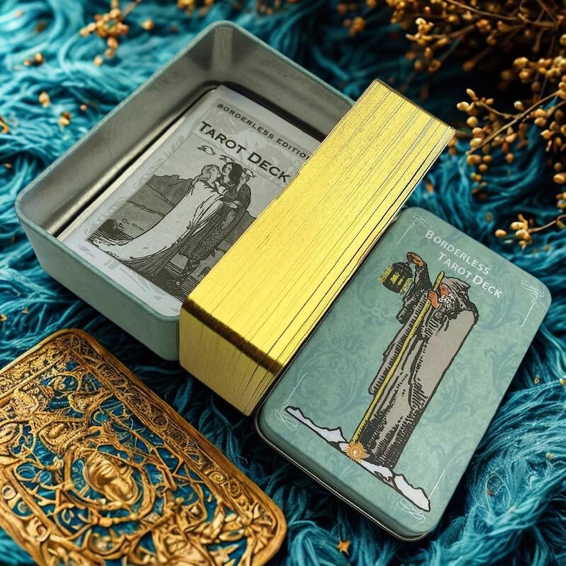 Peut inclure: Un jeu de tarot avec des cartes &agrave; bords dor&eacute;s et une bo&icirc;te en &eacute;tain de couleur sarcelle. La bo&icirc;te porte l'inscription "Borderless Tarot Deck" et pr&eacute;sente des illustrations de personnages. Le jeu est ouvert, r&eacute;v&eacute;lant les cartes et le contenu de la bo&icirc;te.