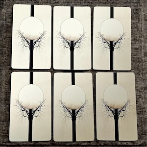 Peut inclure: Six cartes de tarot rectangulaires au design minimaliste. Chaque carte pr&eacute;sente une bande verticale noire, un cercle blanc et une silhouette d'arbre noire. Les cartes sont dispos&eacute;es sur une surface textur&eacute;e de couleur neutre.
