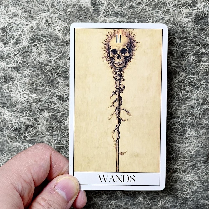 Peut inclure: Une carte de tarot avec un cr&acirc;ne orn&eacute; d'un motif en forme d'&eacute;clat de soleil au sommet d'un b&acirc;ton orn&eacute; de vignes. La carte a un fond cr&egrave;me et le mot "WANDS" en bas.