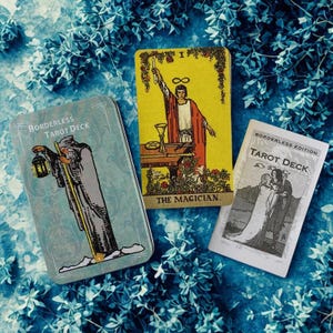 Peut inclure: Un jeu de cartes de tarot avec trois &eacute;l&eacute;ments sur un fond floral bleu. Le jeu comprend une bo&icirc;te en m&eacute;tal avec le texte "BORDERLESS TAROT DECK", une carte jaune avec "THE MAGICIAN" et une carte blanche avec "BORDERLESS EDITION TAROT DECK".