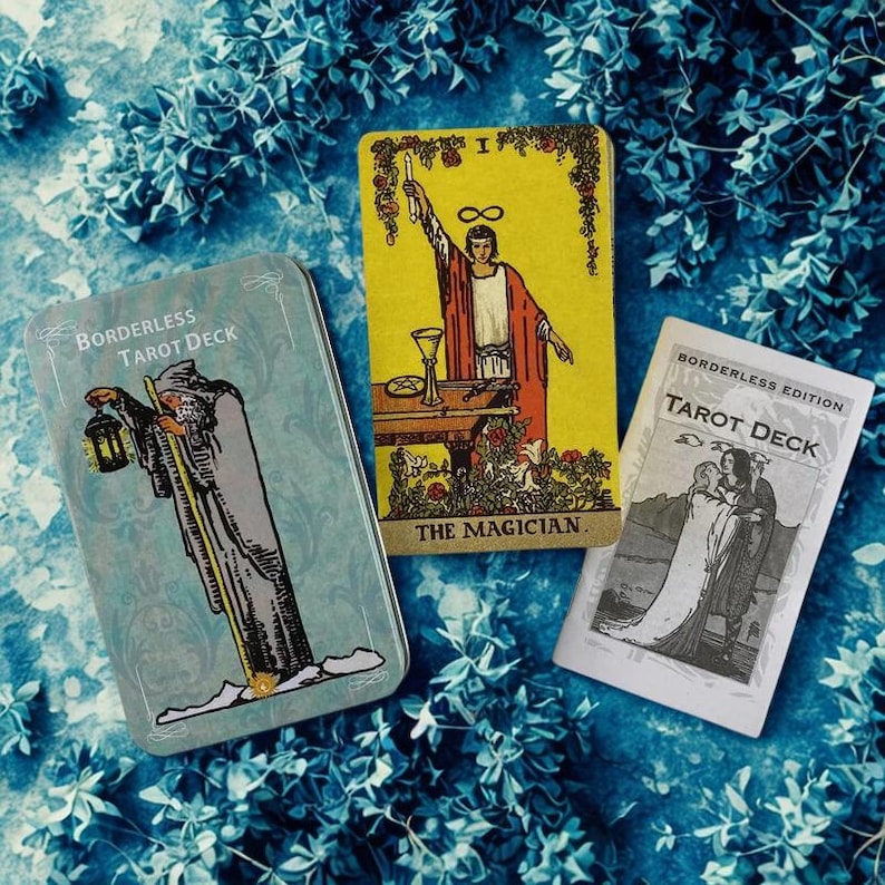 Peut inclure: Un jeu de cartes de tarot avec trois &eacute;l&eacute;ments sur un fond floral bleu. Le jeu comprend une bo&icirc;te en m&eacute;tal avec le texte "BORDERLESS TAROT DECK", une carte jaune avec "THE MAGICIAN" et une carte blanche avec "BORDERLESS EDITION TAROT DECK".