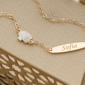 Op de afbeelding: Een goudkleurige kettingarmband met een kleine witte geëmailleerde schaapbedel en een ovaalvormig goudkleurig metalen plaatje met de inscriptie Sofia. De armband is voorzien van een ronde veerringsluiting aan het uiteinde van de fijne ketting.