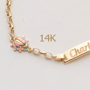 May include: A gold-colored bracelet with a ladybug charm and a rectangular nameplate. The ladybug is pink with black spots and turquoise accents. The nameplate is engraved with the name "Charl". The bracelet is marked "14K".