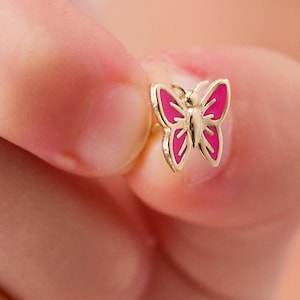 Op de afbeelding: Een goudkleurige vlinder-oorbel met roze emaille details. De vlinder heeft een gedetailleerd ontwerp met een roze omtrek en een goudkleurig lichaam. De oorbel is klein en delicaat, geschikt voor dagelijks gebruik.