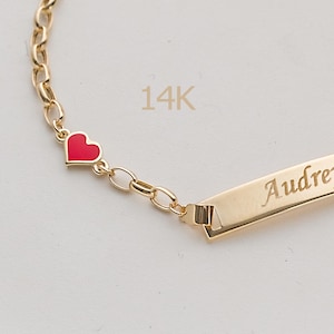 Può includere: Un bracciale a catena in oro 14K con un piccolo ciondolo a forma di cuore in smalto rosso e una barretta rettangolare in oro incisa con il nome Audrey in corsivo. Il bracciale è composto da maglie ovali in oro lucido collegate.