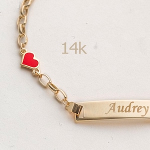 May include: A gold-colored bracelet with a chain and a rectangular nameplate. The name "Audrey" is engraved on the plate. A red heart charm is attached to the chain. The text "14k" is also visible.