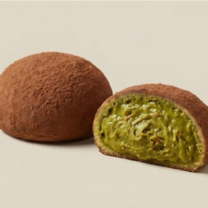 May include: Two round pastries, one whole and one cut in half, sit on a neutral background. The exterior of the pastries is a rich brown, dusted with cocoa powder. The interior of the cut pastry reveals a vibrant green filling.