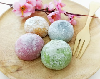 May include: Four colorful mochi desserts on a wooden plate, dusted with powdered sugar. The mochi are pink, green, blue, and tan. Pink cherry blossoms and a wooden fork complete the presentation.