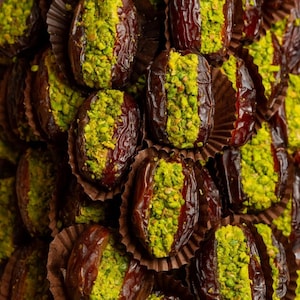 May include: Close-up of a pile of Medjool dates, each halved and filled with bright green pistachio. The dates are dark brown and sit in small brown paper cups. The pistachios provide a vibrant contrast to the dates.
