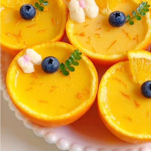 Op de afbeelding: Close-up van sinaasappelhelften gevuld met een geel dessert. Elk dessert is gegarneerd met een blauwe bes, een takje groene bladeren en een witte snoepdecoratie. De desserts zijn gerangschikt op een wit bord met geschulpte rand.