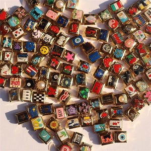May include: A selection of gold-coloured rectangular charms with diverse, colourful designs. The charms display images such as a smiley face, a strawberry, a black cat, and the word "sexy". The charms are arranged on a white surface.
