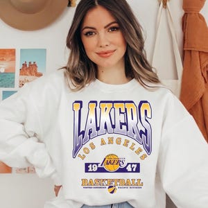 May include: A white crewneck sweatshirt featuring a graphic print that reads LAKERS LOS ANGELES 1947 BASKETBALL with a basketball icon and team colours in purple, gold, and white. The design includes the text WESTERN CONFERENCE PACIFIC DIVISION.
