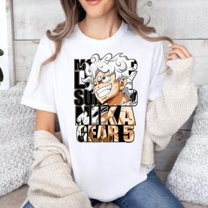 May include: White t-shirt featuring a colorful anime graphic. The graphic includes a character with white hair and the text "MONKEY D. LUFFY SUN NIKA GEAR 5" in a bold, black font. The shirt is a casual, everyday wear item.