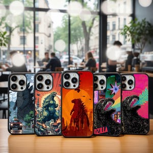 May include: A collection of phone cases with various Godzilla-themed designs. The cases feature illustrations of Godzilla in different poses, set against cityscapes and colorful backgrounds. The cases are displayed on a wooden surface.