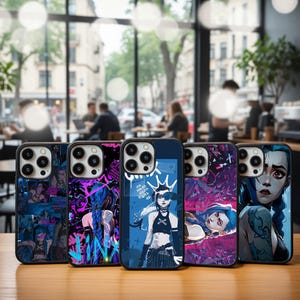 May include: A collection of five phone cases featuring colorful illustrations. The cases showcase various character designs with a blue and purple color scheme. The cases are displayed on a wooden surface with a blurred cafe background.