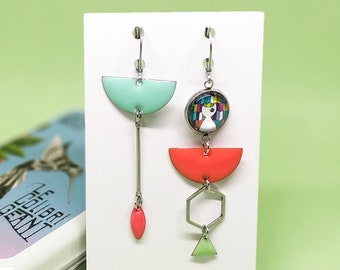 May include: Geometric dangle earrings with mint green, coral, and silver-tone metal elements. One earring features a long bar with a coral teardrop, while the other has a cat design and a geometric shape.