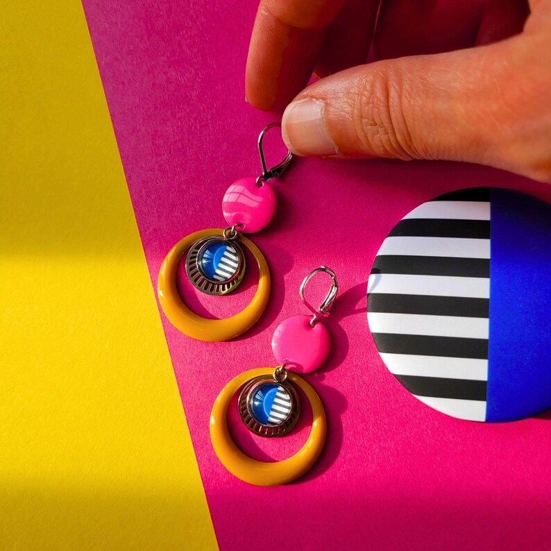 May include: A pair of vibrant earrings with a retro design. The earrings feature a yellow hoop, a pink bead, and a circular pendant with a blue and white striped design. A matching round pin with black and white stripes and a blue section is also visible.