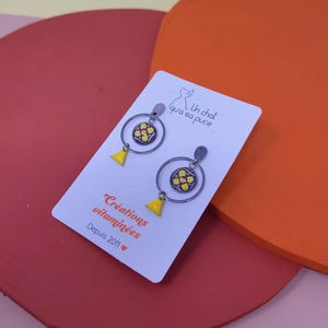 K&ouml;nnte beinhalten: Ein Paar gelbe und orangefarbene Blumen-Hoop-Ohrringe mit einem kleinen Dreiecksanh&auml;nger, der an jedem Reifen h&auml;ngt. Die Ohrringe befinden sich auf einer wei&szlig;en Karte mit dem Text "Un chat qui a sa puce." und "Cr&eacute;ations vitamin&eacute;es Depuis 2011".