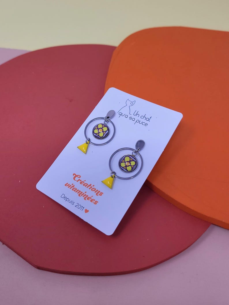 K&ouml;nnte beinhalten: Ein Paar gelbe und orangefarbene Blumen-Hoop-Ohrringe mit einem kleinen Dreiecksanh&auml;nger, der an jedem Reifen h&auml;ngt. Die Ohrringe befinden sich auf einer wei&szlig;en Karte mit dem Text "Un chat qui a sa puce." und "Cr&eacute;ations vitamin&eacute;es Depuis 2011".