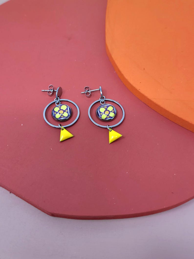 K&ouml;nnte beinhalten: Ein Paar silberne Ohrringe mit einem gelben und blauen Blumendesign. Die Ohrringe haben eine runde Form mit einem dreieckigen Anh&auml;nger.