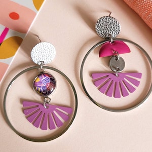 May include: A pair of silver-tone hoop earrings with a mix of geometric and floral designs. Each earring features a textured silver disc, a circular floral pendant, and a fan-shaped magenta accent. The earrings are set against a pink background.