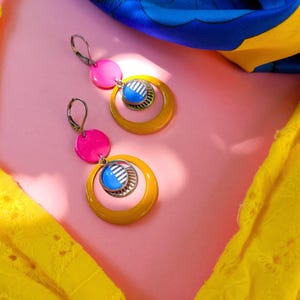 May include: A pair of vibrant earrings with a retro design. Each earring features a bright pink disc, a mustard yellow hoop, and a blue and silver circular pendant. The earrings are set against a pink background with yellow and blue fabric accents.