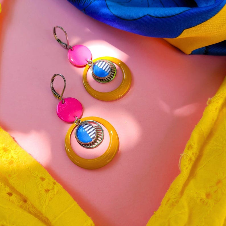 May include: A pair of vibrant earrings with a retro design. Each earring features a bright pink disc, a mustard yellow hoop, and a blue and silver circular pendant. The earrings are set against a pink background with yellow and blue fabric accents.
