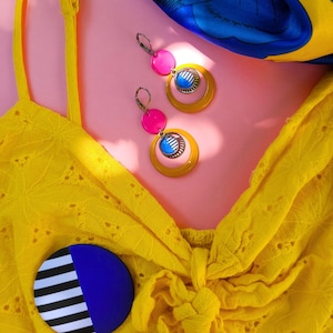 May include: A pair of colorful earrings with pink, yellow, and blue accents. The earrings are paired with a yellow top and a blue and yellow scarf on a pink background. A round pin with a blue and white design is also visible.