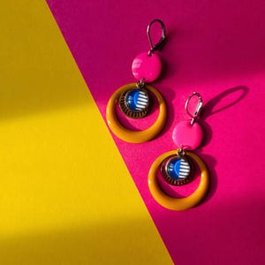 May include: A pair of vibrant earrings with a retro design. Each earring features a bright pink disc, a mustard yellow hoop, and a smaller circular pendant with blue and white stripes. The earrings are set against a pink and yellow background.