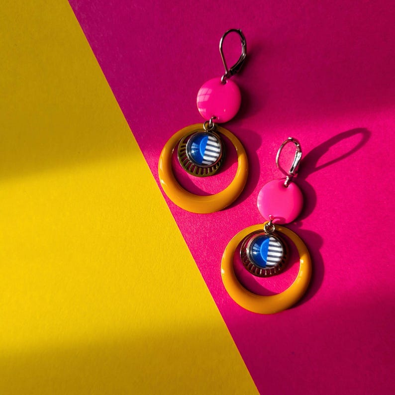 May include: A pair of vibrant earrings with a retro design. Each earring features a bright pink disc, a mustard yellow hoop, and a smaller circular pendant with blue and white stripes. The earrings are set against a pink and yellow background.