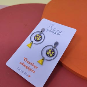 K&ouml;nnte beinhalten: Ein Paar silberne Creolen mit einem gelben und violetten Blumenanh&auml;nger und einem gelben Dreiecksanh&auml;nger. Die Ohrringe werden auf einer wei&szlig;en Karte mit dem Text "Un chat qui a sa puce" und "Cr&eacute;ations vitamin&eacute;es Depuis 2011" pr&auml;sentiert.