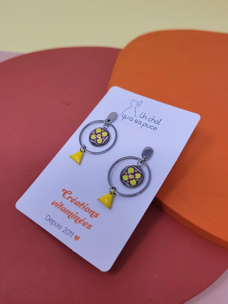 K&ouml;nnte beinhalten: Ein Paar silberne Creolen mit einem gelben und violetten Blumenanh&auml;nger und einem gelben Dreiecksanh&auml;nger. Die Ohrringe werden auf einer wei&szlig;en Karte mit dem Text "Un chat qui a sa puce" und "Cr&eacute;ations vitamin&eacute;es Depuis 2011" pr&auml;sentiert.
