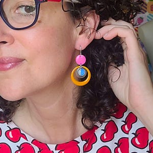 May include: A pair of colorful earrings with a pink circle, a yellow hoop, and a blue and silver accent. The earrings are hanging from a silver hook. The earrings are a fun and unique accessory.