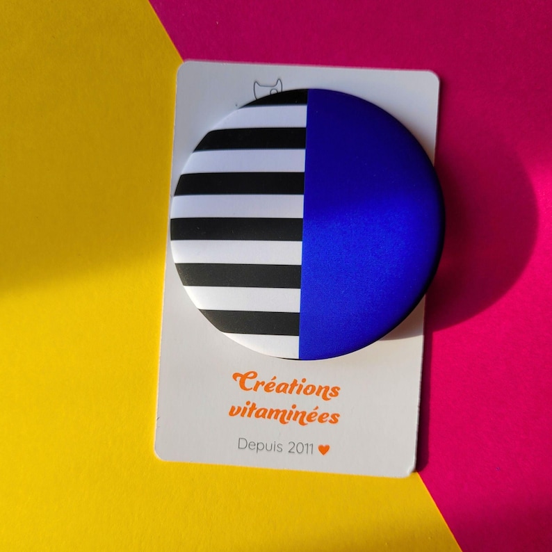 May include: A round button with a split design: one half features black and white horizontal stripes, the other half is solid blue. The button is on a white card with the text "Cr&eacute;ations vitamin&eacute;es Depuis 2011". The background is yellow and pink.