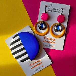 May include: A pair of earrings and a round pin with a geometric design. The earrings have pink and yellow accents with a circular design. The pin has a blue and black and white striped design. The background is yellow and pink.