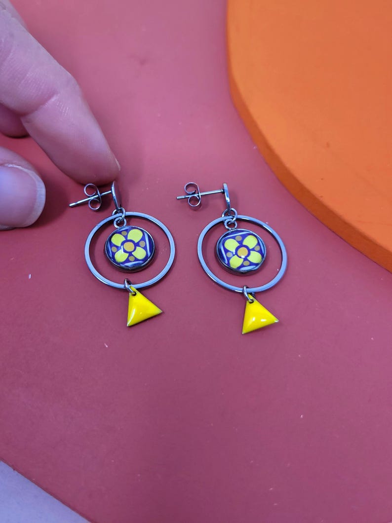 K&ouml;nnte beinhalten: Ein Paar silberne Creolen mit einem gelben und blauen Blumenmuster und einem gelben Dreiecksanh&auml;nger, der an jeder Creole h&auml;ngt.