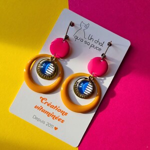 May include: A pair of handmade earrings with a bright color scheme. The earrings feature a hot pink disc, a mustard yellow hoop, and a circular pendant with a blue and white design. The earrings are displayed on a white card with text and a cat illustration.