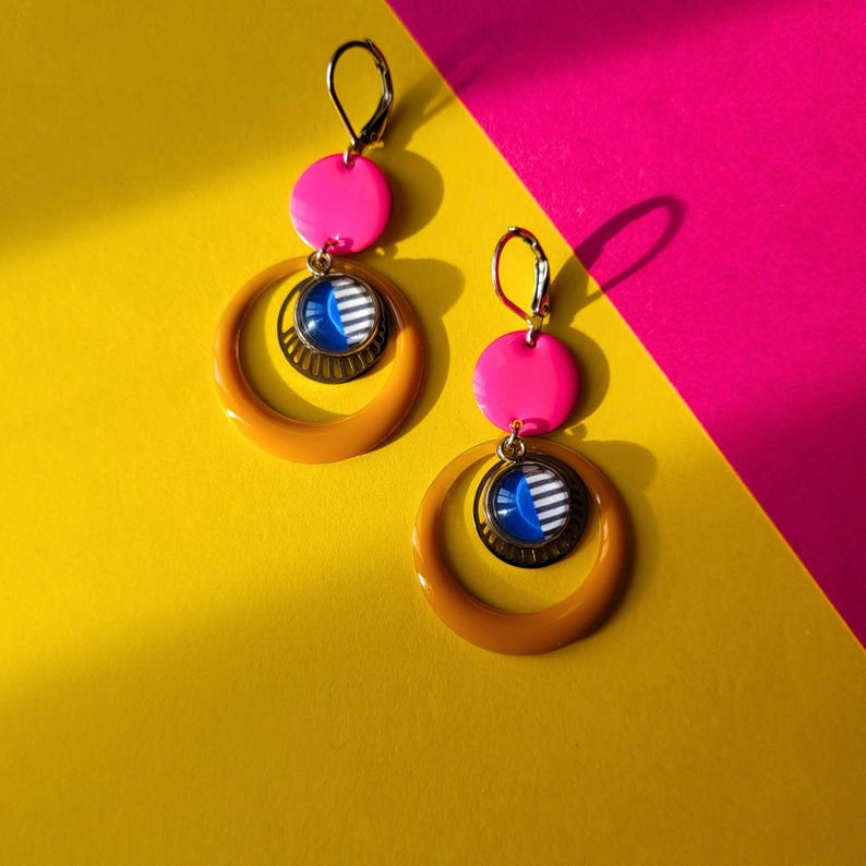 May include: A pair of vibrant earrings with a retro design. Each earring features a bright pink disc, a circular pendant with blue and white stripes, and a large, orange hoop. The earrings are set against a yellow and pink background.